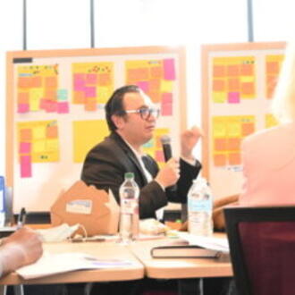 Two people seated, post it notes suggesting workshopping in background