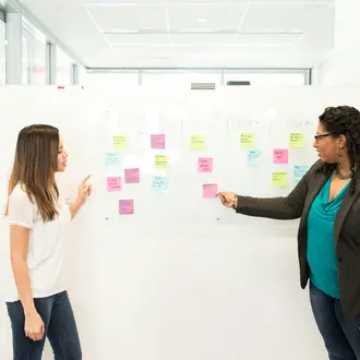 Two women standing in front of a whiteboard with sticky notes on it