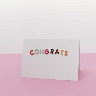 A white card with the word "congrats" on it in colorful letters, against a pink-and-white background