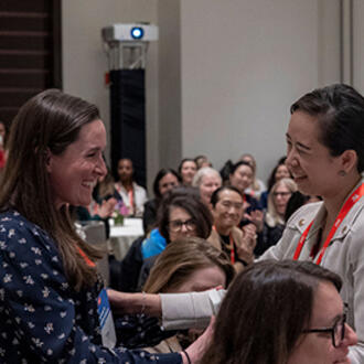 Two women embrace in the foreground of a packed room at a conference