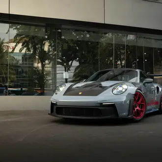Photo of silver Porsche with a black hood and red rims parked outside of a glass storefront.
