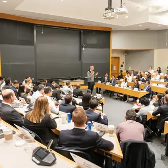 Wide shot of a classroom filled with students facing professor lecturing up front. Behind the professor are empty blackboards.