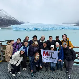 Study tour attendees pose in a group photo in front of a glacier holding an MIT sign