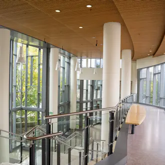 Empty second floor hallway in e62. To the left is a staircase down to the first floor. To the right is the bridge that leads to building E52 if you keep walking straight. There is a woman out of focus walking on the bridge towards the camera all the way to the right of the bridge.
