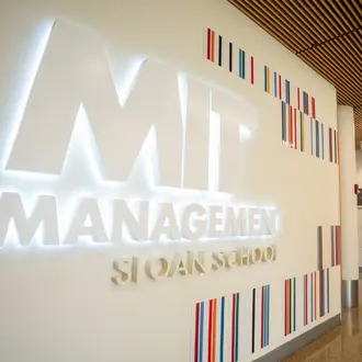 Illuminated MIT Sloan School of Management sign in a campus hallway, with a blurred person walking past.