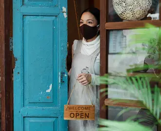 Woman in a face mask opening the door of a business holding a "Welcome, we're open" sign looking tentative and unsure.