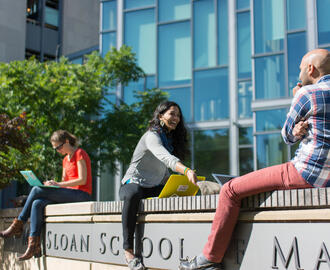 Three young adults sit on a wall outside the MIT Sloan School of Management
