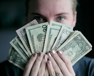Image of the face of a woman who is holding U.S. currency fanned out in her hands, obscuring the lower half of her face. 