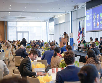 Wide photograph of conference with many people sitting at tables listening to presentations