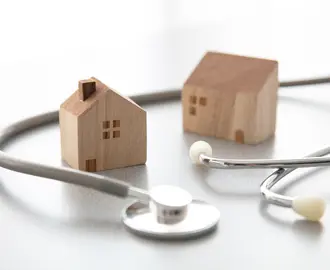 Two mini wooden house figurines with a regular sized stethoscope wrapped around them on smooth white surface