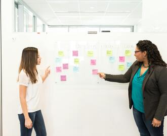 Two women standing in front of a whiteboard with sticky notes on it