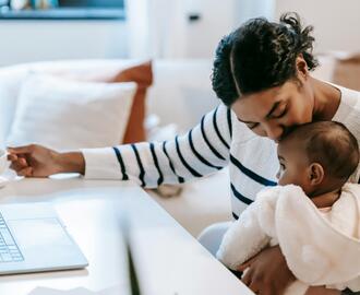 Image of a woman holding a baby in one arm and sitting near a computer