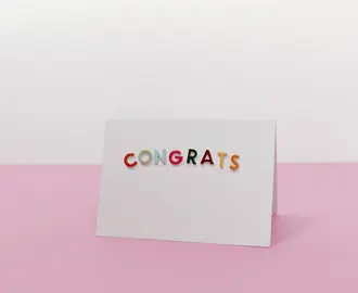 A white card with the word "congrats" on it in colorful letters, against a pink-and-white background