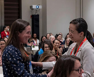 Two women embrace in the foreground of a packed room at a conference