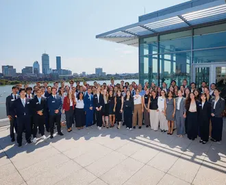 The MBAn Class of 2025 lines up for a photo outside with the Charles River and the Boston skyline in the background