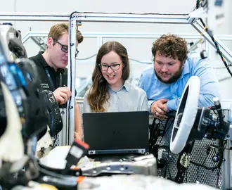 Three people look at a laptop in a some type of industrial setting