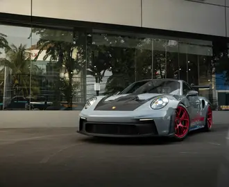 Photo of silver Porsche with a black hood and red rims parked outside of a glass storefront.