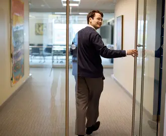 PhD student Alex Busch holds open a door and smiles over his shoulder as he walks through a building at MIT Sloan