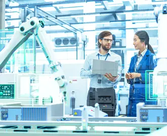 Employees in discussion inside a manufacturing plant with robotic arms and digital screens