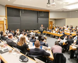 Wide shot of a classroom filled with students facing professor lecturing up front. Behind the professor are empty blackboards.