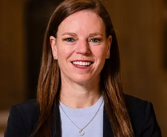 Photo of Megan Greene, a fair-skinned woman with long brown hair and light eyes wearing a dark blazer, a light blue sweater, and a simple necklace, smiling.