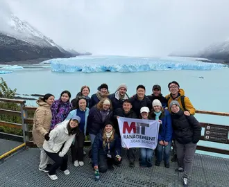 Study tour attendees pose in a group photo in front of a glacier holding an MIT sign