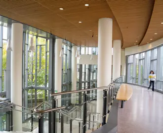 Empty second floor hallway in e62. To the left is a staircase down to the first floor. To the right is the bridge that leads to building E52 if you keep walking straight. There is a woman out of focus walking on the bridge towards the camera all the way to the right of the bridge.