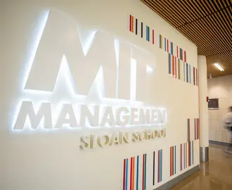 Illuminated MIT Sloan School of Management sign in a campus hallway, with a blurred person walking past.