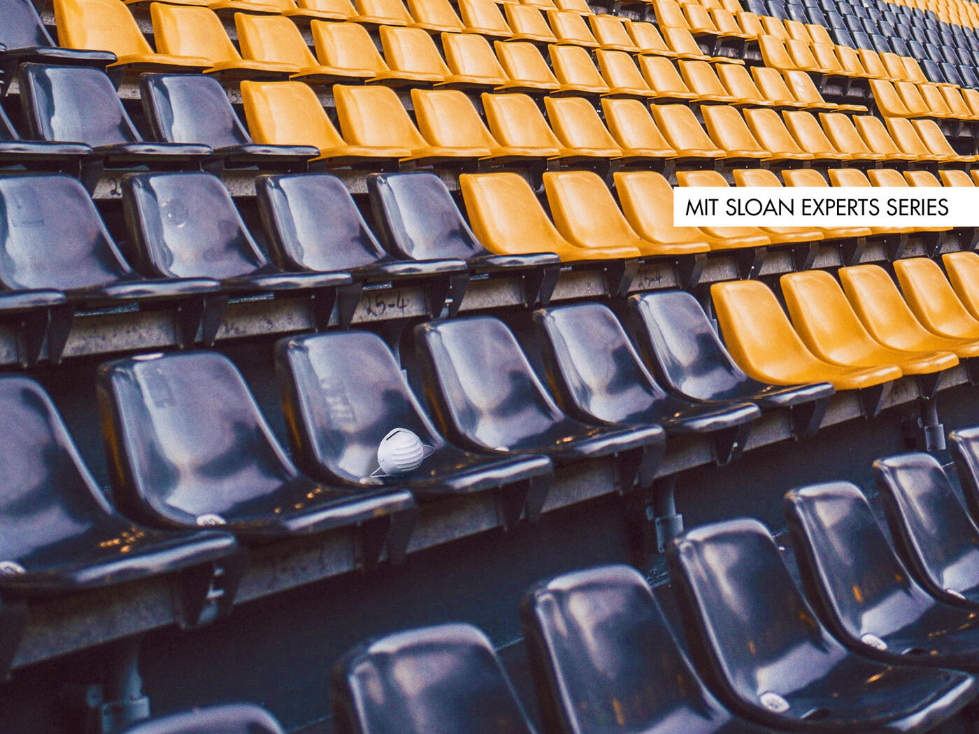 Photograph of empty stadium seats. One seat has a face mask on it.