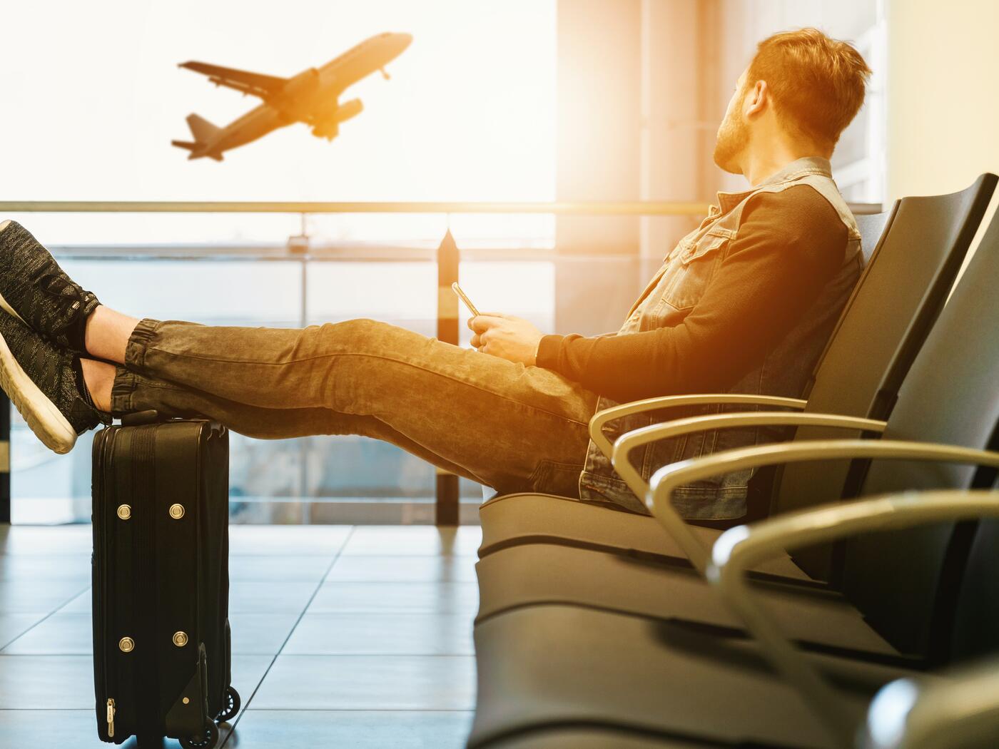 Man sitting in airport waiting area, staring out window at a plane taking off
