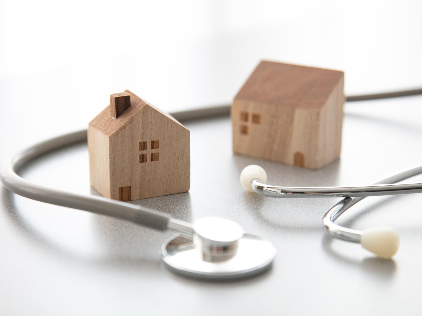Two mini wooden house figurines with a regular sized stethoscope wrapped around them on smooth white surface