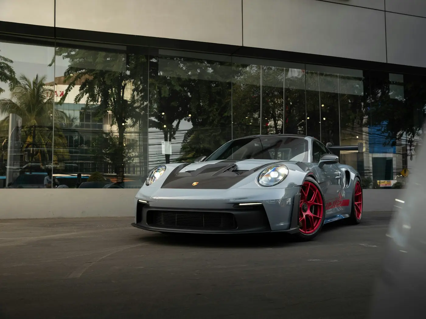 Photo of silver Porsche with a black hood and red rims parked outside of a glass storefront.