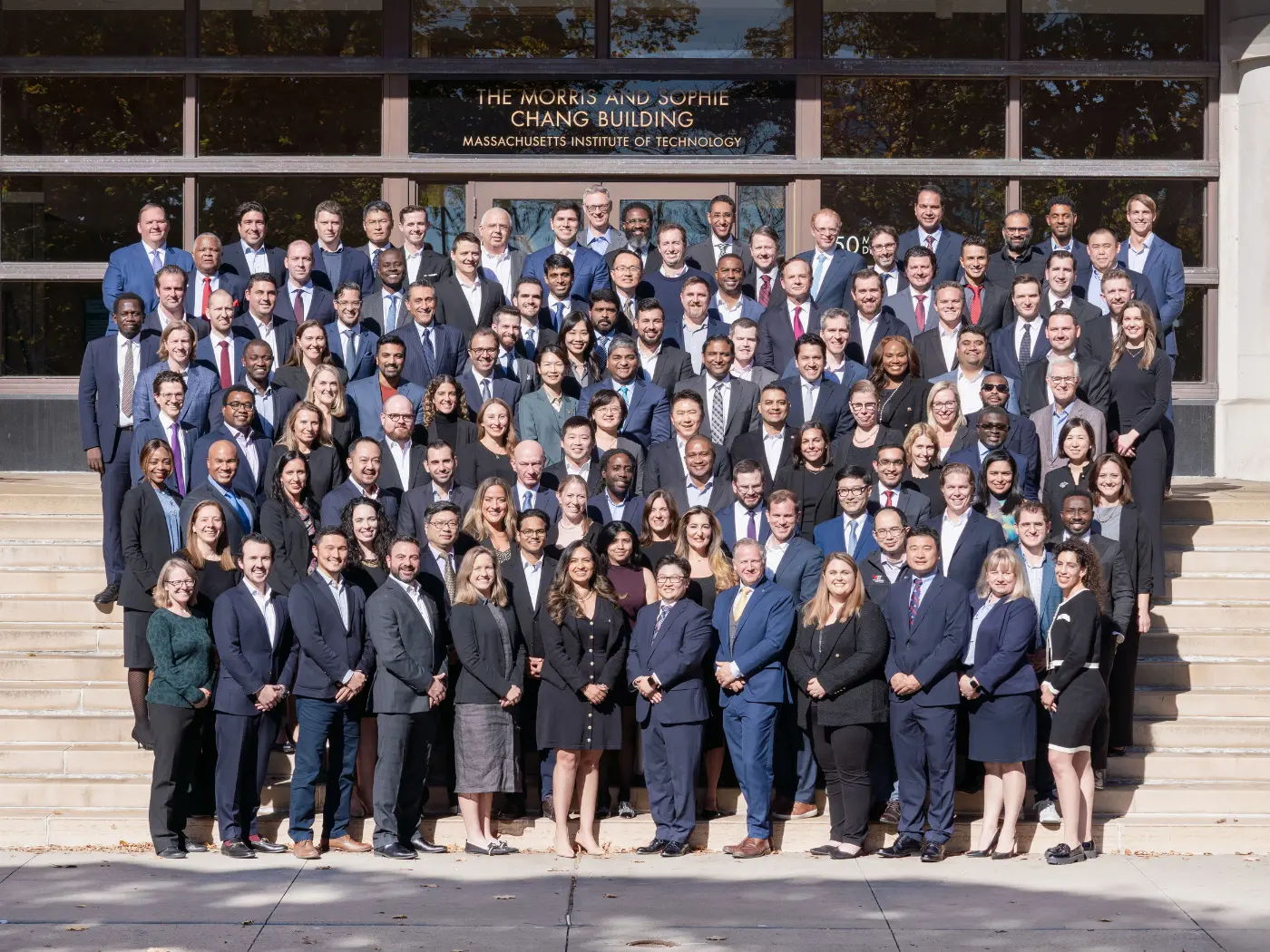 MIT EMBA Class of 2026 standing on the stairs in front of the Memorial Drive Entrance to E52. They are wearing dark business attire.