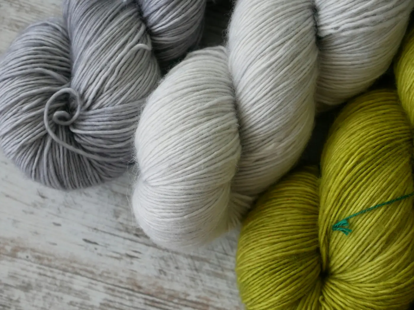 Photo of 3 skeins of yarn: 1 silvery gray, 1 white, and 1 leaf green, sitting on a gray wooden surface