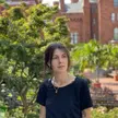 A photo of Guila Crippa wearing a black top, looking away from the camera, outside in front of a background of trees and brick buildings
