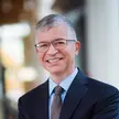 Photo of a person with grey hair wearing a dark suit, a blue shirt, a tie, and glasses