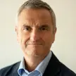 Photo of a person with short grey hair wearing a blue shirt and dark jacket