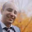 Man wearing a gray suit, light shirt, and blue striped tie, against a blurred background of a tree with yellow leaves.