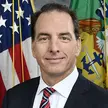 Photo of a man wearing a navy  blue suit, white shirt, and red-white-and-blue-striped tie. Backdrop is a US flag and a US Marine Corps flag