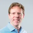 photo of a man with short hair and glasses wearing a blue shirt against a gray  background