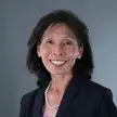 Woman with dark hair wearing a dark suit, lilac colored blouse, and pearl earrings and necklace.