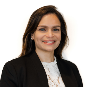 A headshot of Neha in front of a white background. Neha is a woman with brown hair, smiling, and wearing a white shirt under a black blzaer