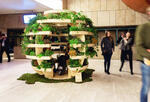 a large wooden sphere with green plants on it sits in a large company lobby with people walking by
