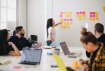 People having a meeting around a table, standing woman pointing to sticky notes on wall